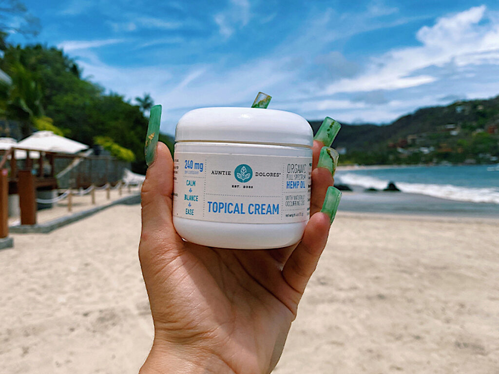 Image of Auntie Dolores Topical Cream canister with the beach in the background